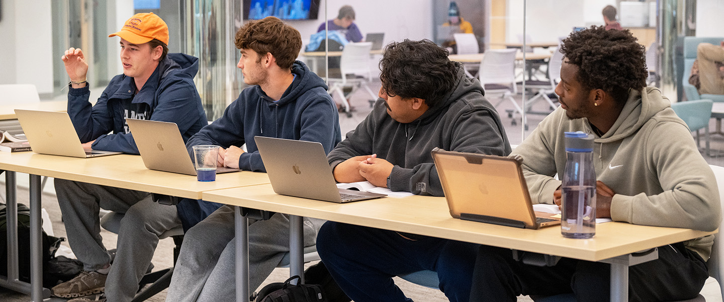 Students sitting in a row at a table with laptops open listening to another student at Hampden-Sydney College