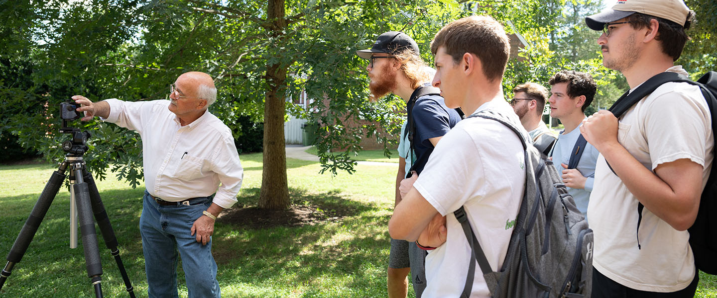 Art students standing outdoors watching their professor look through a camera