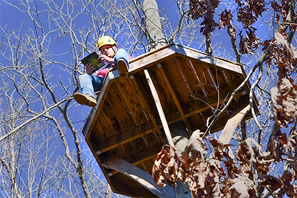 Jack Garst '21 sitting atop a high ropes platform with a book