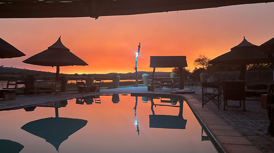 a pool surrounded by huts with the sunset in the distance