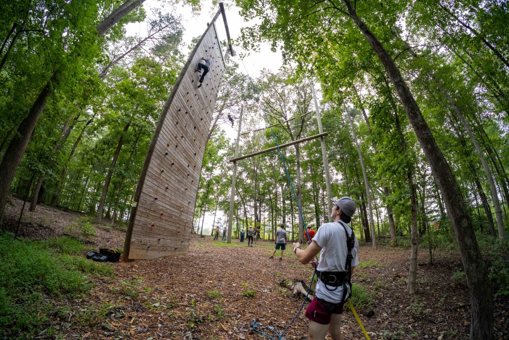 Students surrounding a climbing wall at an outdoor ropes course